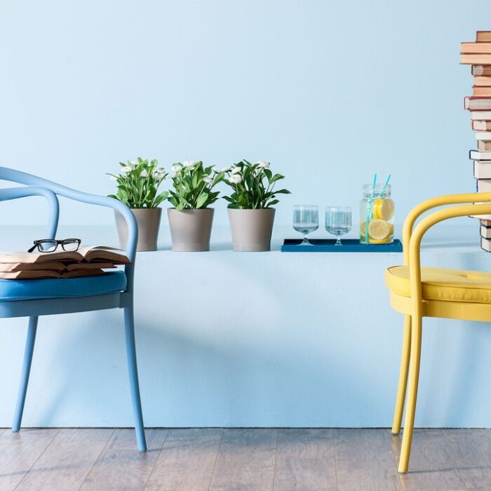 A serene reading nook featuring a cool blue wall, two vibrant chairs in shades of blue and yellow, a stack of books teetering towards the ceiling, a refreshing pitcher of lemon water, and potted green plants adding a touch of nature.
