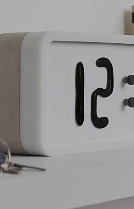 A modern digital clock displaying the time "12:00" with a minimalist design, sitting on a bedside table next to a set of keys.