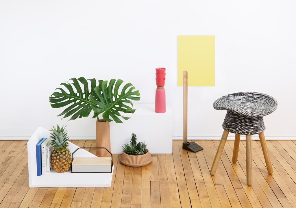 A bright and minimalist living space featuring modern decor with a monstera plant, stack of books with a pineapple on top, two pink vases, a yellow canvas, and a unique patterned stool.