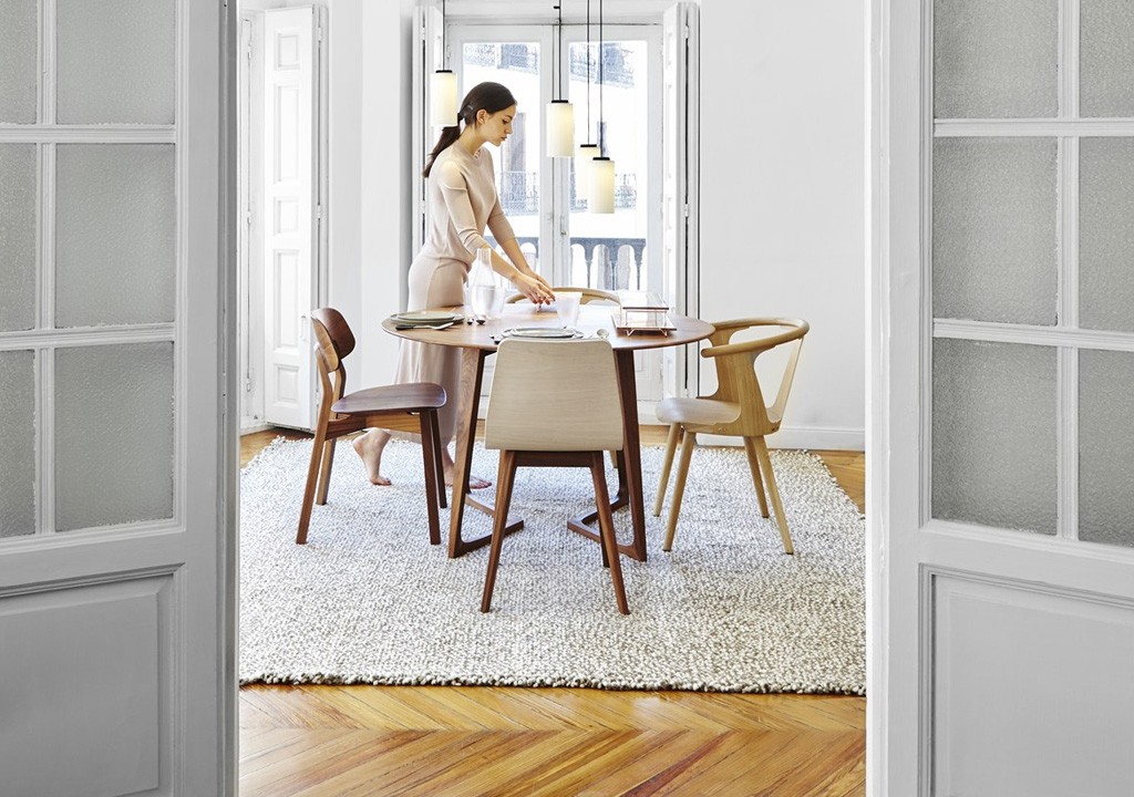 A woman in a sleek outfit sets a modern dining table in a bright, stylish room with open french doors.
