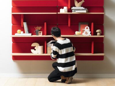 A person in a striped sweater crouches while organizing items on a vibrant red shelving unit against a white wall.