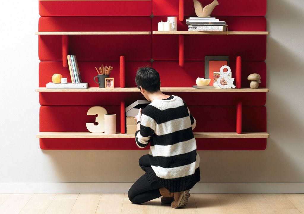 A person in a striped sweater crouches while organizing items on a vibrant red shelving unit against a white wall.
