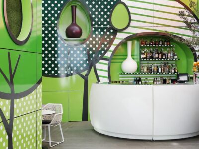 A vibrant and modern bar area with a whimsical forest theme, featuring a white curved bar counter, stool seating, and a wall mural with abstract trees and dotted patterns.